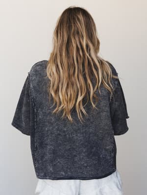 A back-view shot of a boxy, acid-washed charcoal tee with raw-edge finishing — the kind of top that looks like it's been lived in for years in the best possible way.