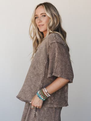 A relaxed, washed mocha oversized top styled with a stack of colorful beaded bracelets and silver rings — the kind of effortless outfit that looks like you tried just enough.