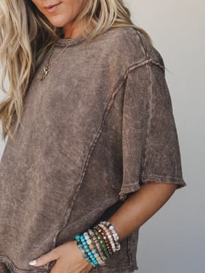 A close-up lifestyle shot of a washed mocha textured tee styled with a stack of colorful beaded bracelets and a dainty gold charm necklace — the kind of effortless layering that looks accidental but absolutely isn't.