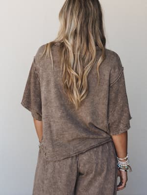 A back-view shot of a matching mineral-washed mocha set — a relaxed cropped top with raw edge seam details paired with wide-leg pants, styled with stacked boho bracelets for an effortlessly lived-in feel.