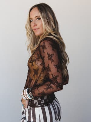 A model shot in three-quarter profile wearing a sheer brown lace floral long sleeve top layered over a mustard underlayer, paired with wide-leg striped pants and a stack of mixed beaded bracelets — the kind of outfit that looks intentional without tr