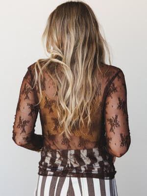 Shot from behind, this sheer brown lace top layers over what looks like a warm amber-toned underlayer, its floral mesh pattern catching soft studio light while wavy blonde hair and a stripe-print bottom round out the effortlessly undone feel.