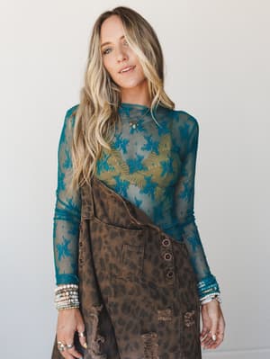 A sheer teal floral lace long-sleeve top layered over an olive cami, styled with a distressed leopard print overall skirt and stacked boho bracelets — the kind of outfit that feels vintage-collected and completely personal.