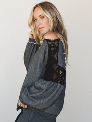 A relaxed off-shoulder sweatshirt top in charcoal gets an unexpected boho twist with a bold black crochet lace panel running down the back — the kind of piece that looks like a lazy Sunday until you turn around.