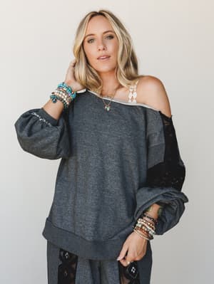 An oversized charcoal sweatshirt top with unexpected lace paneling gets styled off-the-shoulder with stacked boho bracelets and a delicate pendant necklace — the kind of effortless outfit that looks like you tried just the right amount.
