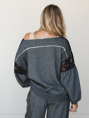 Shot from the back, a relaxed charcoal sweatshirt top with black crochet lace insets at the sleeves and exposed raw-edge seam detailing down the back — styled with matching wide-leg pants for an effortlessly put-together, boho-comfortable look.