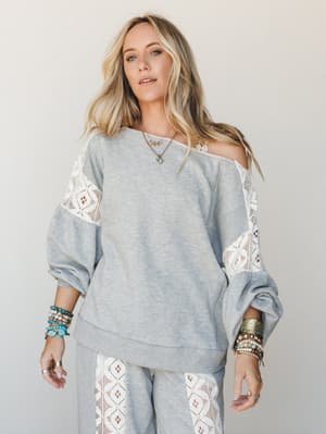 A relaxed heathered grey sweatshirt top with geometric lace inset panels styled off-the-shoulder, layered with turquoise and gold jewelry for an effortlessly boho everyday look.