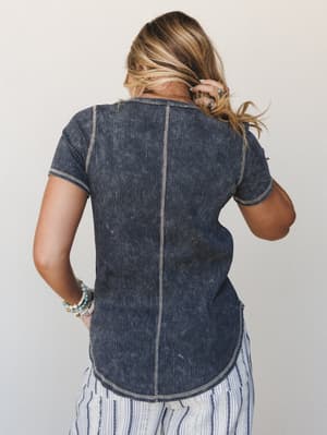 Shot from the back, a model in a mineral-washed ribbed charcoal tee with visible contrast stitching and a curved hem — styled with striped wide-leg pants and stacked beaded bracelets for that effortlessly collected, off-duty feel.
