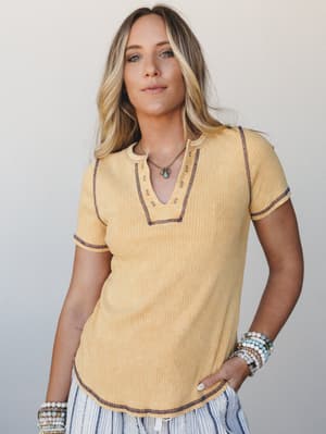 A ribbed mustard yellow henley tee with contrast navy stitching and a notched neckline, styled with stacked stone bracelets and a turquoise pendant necklace — the kind of effortless top that looks just as good tucked into jeans as it does thrown on b