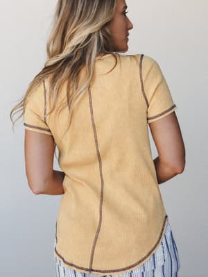 A back-view shot of a ribbed mustard tee with contrast navy stitching along the seams and hem, paired with blue striped wide-leg pants — casual and collected with just enough detail to keep it interesting.