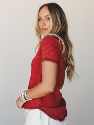A relaxed ribbed short-sleeve tee in a rich crimson-rust, shot in profile against a soft studio wall — the kind of top that looks just as good tucked into jeans as it does thrown on over pajama pants.