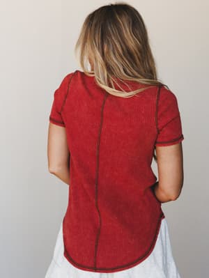 A back-facing shot of a ribbed rust tee with visible contrast stitching and a center seam — the kind of top that looks effortless tucked into a white skirt or worn straight through the weekend.