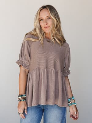 A relaxed ribbed peplum top in warm acorn brown styled with jeans and stacked turquoise and beaded bracelets — the kind of effortless, lived-in look that works as hard as you do without trying to prove it.