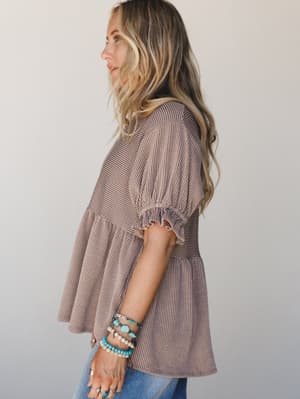 A tiered babydoll top in warm acorn brown with a fine stripe texture, styled with stacked turquoise and beaded bracelets and a peek of blue denim — casual and expressive with a relaxed boho ease.
