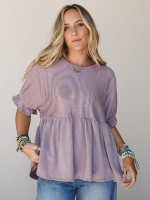A relaxed peplum top in soft orchid purple with a subtle ribbed stripe texture, styled with wide-leg denim and stacked turquoise bracelets — the kind of top that makes a Tuesday feel a little more intentional.