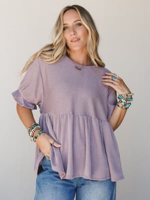 A relaxed ribbed peplum top in dusty orchid worn with jeans and stacked turquoise and beaded bracelets — the kind of top that looks effortless but clearly has a point of view.