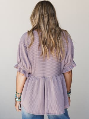 A back-view shot of a soft orchid ribbed peplum top with ruffle-cuffed sleeves, styled with jeans and stacked turquoise bracelets — relaxed and pretty in equal measure.