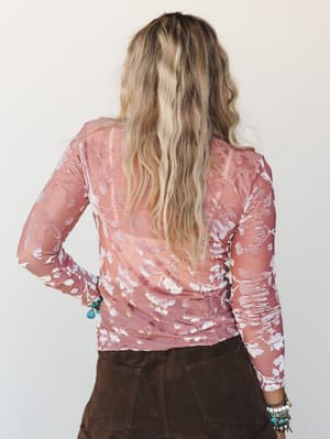 Shot from behind, a model in a sheer dusty rose floral mesh long-sleeve top paired with a dark chocolate mini skirt — the kind of layering piece that makes everything feel a little more intentional.