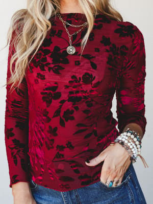 A rich wine-red burnout velvet floral top styled with layered gold necklaces, stacked beaded bracelets, and silver rings — the kind of top that makes jeans feel like a whole moment.