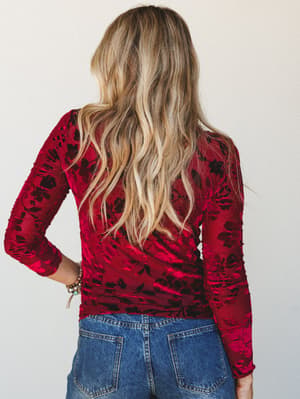 Shot from behind, a model in a rich wine-red burnout velvet floral long sleeve top paired with light-wash denim — the kind of top that makes a Saturday night feel intentional.