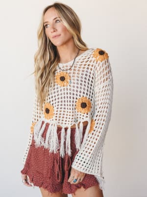 A cream open-weave crochet top with sunflower motifs and fringe hem paired with dusty rose micro shorts — the kind of outfit that looks effortless but was absolutely chosen on purpose.
