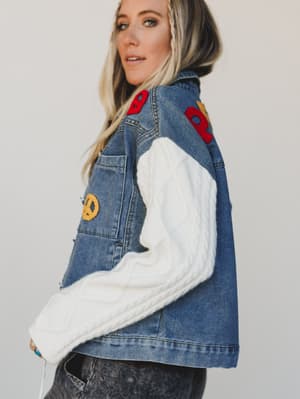 Person wearing a denim jacket with colorful patches and a white sleeve against a plain background