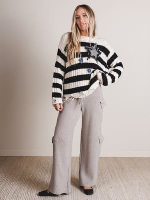 Woman wearing a black and white striped sweater with star patterns and gray pants against a plain background
