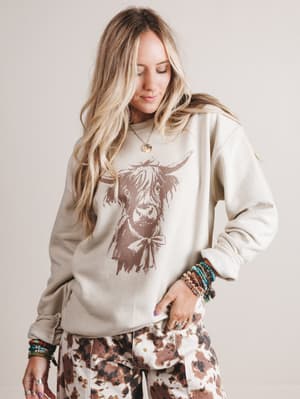 Woman wearing a beige sweatshirt with a cow graphic and patterned pants on a plain background
