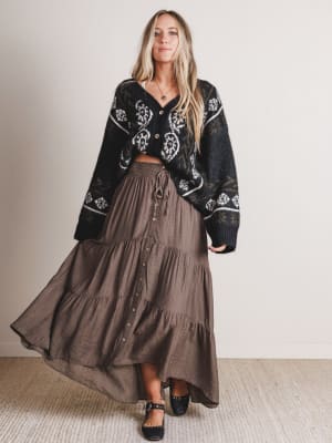 Woman wearing a black embroidered cardigan and brown tiered skirt against a plain background