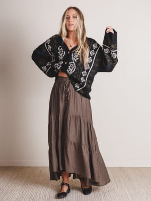 Woman wearing a black embroidered cardigan and brown tiered skirt against a plain background