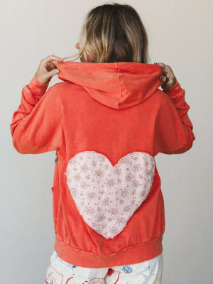 A model shown from the back wearing a washed coral hoodie with a large floral-print heart appliqué centered on the back — the kind of piece that makes people stop you on the street to ask where you got it.
