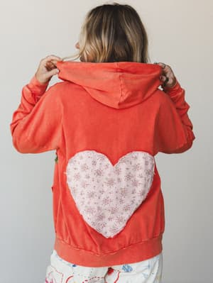 A model shown from the back wearing a washed coral hoodie with a large floral-print heart appliqué centered on the back — the kind of piece that makes people stop you on the street to ask where you got it.
