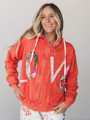 A smiling model wears a washed coral zip-up hoodie with patchwork 'LOVE' letter appliqués in mixed prints, styled over printed white pants — playful, warm, and totally the kind of jacket you'd wear on a coffee run and get compliments on.
