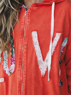 A close-up shot of a washed orange zip-up hoodie with a raw-edge fabric letter 'V' appliqué on the chest and patchwork fabric details along the sides — the kind of piece that feels personal before you've even worn it.