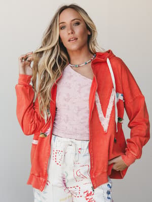 A relaxed, color-happy outfit built around a washed coral zip-up hoodie with a lace chevron appliqué — layered over a pink lace top and printed drawstring pants, with a beaded pearl necklace tying it all together in the most effortlessly playful way.
