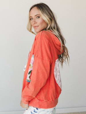 A model shot from the side showing a washed coral-orange hoodie jacket with a patchwork graphic on the front and a floral fabric pocket detail on the back — the kind of piece that looks like it has a whole personality before you even put it on.