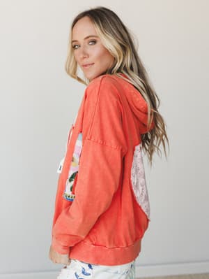 A model shot from the side showing a washed coral-orange hoodie jacket with a patchwork graphic on the front and a floral fabric pocket detail on the back — the kind of piece that looks like it has a whole personality before you even put it on.