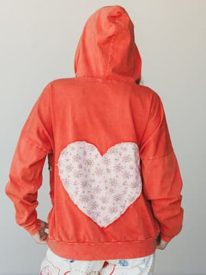 The back of a washed coral hoodie sweatshirt with a large heart-shaped floral fabric patch appliquéd at center back — the kind of piece that feels personal, like someone made it just for you.