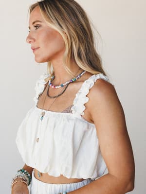 A cream cropped top with ruffled straps and button-front detail shot in soft profile — layered with beaded necklaces and stacked bracelets, the whole look has that easy, jewelry-box-spilled-over energy.