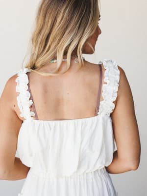 A close-up back view of a cream textured top with ruffle-trimmed straps and an open back, styled with layered delicate necklaces — soft, romantic, and effortlessly warm-weather ready.