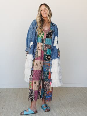 A full-length model shot of a layered boho look — an oversized acid-wash denim jacket with tiered white ruffle sleeves and floral appliqué worn open over a mixed patchwork maxi dress, finished with sky blue buckle sandals.