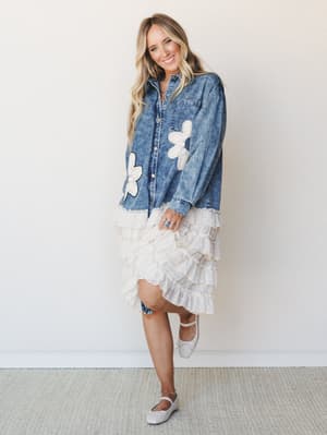 A washed indigo denim jacket with oversized daisy appliqués is styled over a cream tiered ruffle midi skirt, giving off that effortless mix of vintage denim and romantic femininity.