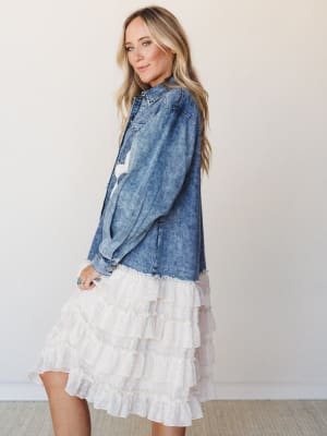 A breezy denim jacket with a surprising ruffle trim finish is styled over a cream tiered skirt, shot in soft studio light — the kind of outfit that makes a bridal shower feel effortlessly personal.