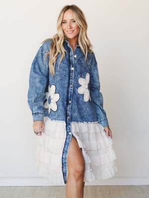 A denim shirt-jacket with oversized crochet daisy appliqués transitions into a tiered cream ruffle hem — equal parts playful and unexpected, styled simply against a soft studio wall.