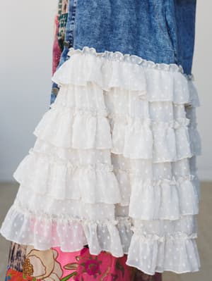 A close-up detail shot of a washed indigo denim jacket with a dramatic tiered ruffle hem in cream swiss dot fabric, layered over a bright floral skirt — the contrast between structured denim and soft, romantic ruffles is the whole story.