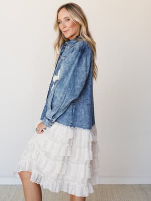 A breezy denim jacket with a surprising ruffle trim finish is styled over a cream tiered skirt, shot in soft studio light — the kind of outfit that makes a bridal shower feel effortlessly personal.