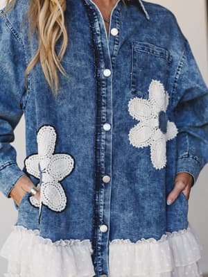 A close-up of a washed indigo denim jacket layered over a white ruffle hem dress, with oversize crochet flower appliqués on the front — the kind of piece that looks like it has a personality before you even put it on.