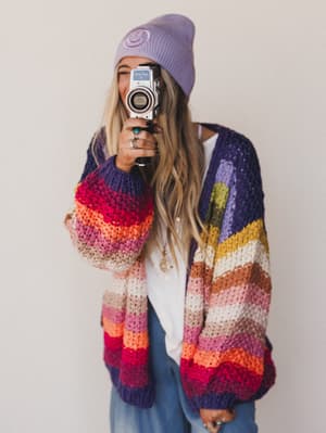 A woman with long blonde hair wears a purple beanie and the Wrapped up in Color Knit Cardigan - Rainbow as she smiles, holding a vintage film camera to her face against a plain white background.