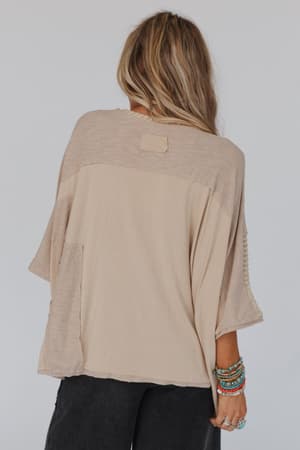 Wearing the City Drifter Oversized Contrast Top in taupe, a woman with long blonde hair faces away. She embraces boho style with colorful beaded bracelets on her right wrist and dark pants.