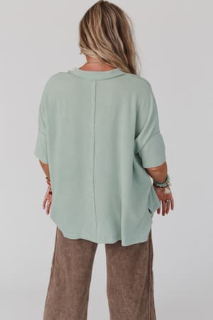 A person with long, wavy hair, wearing The Nest On The Go V Neck Ribbed Essential Tee in Sage from Three Bird Nest and loose brown pants, stands facing away from the camera against a plain background.